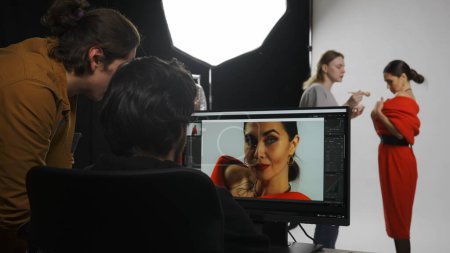 Photo for Photo production and creative teamwork concept. Backstage of model and professional team in the studio. Editor and photographer talking checking photos looking at monitor, assistant fixes makeup. - Royalty Free Image