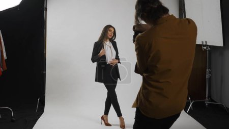 Photo for Photo production and creative teamwork concept. Backstage of model and professional team in the studio. Full shot attractive female model in suit posing, photographer taking pics, serious expression. - Royalty Free Image