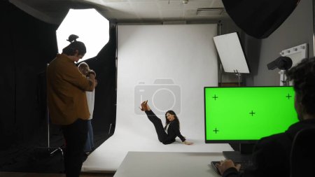 Photo for Photo production and creative teamwork concept. Backstage of model and professional team in the studio. Full shot appealing model in suit posing on the ground, editor monitor chroma key green screen. - Royalty Free Image