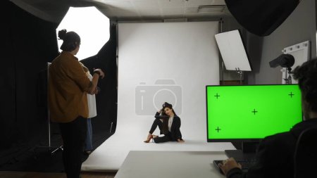 Photo for Photo production and creative teamwork concept. Backstage of model and professional team in the studio. Full shot female model in suit posing on the floor, editor monitor chroma key green screen. - Royalty Free Image