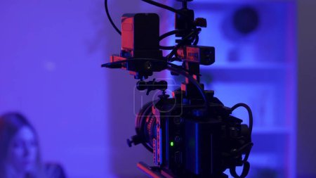 Téléchargez les photos : Caméra vidéo professionnelle sur trépied sur fond flou d'une présentatrice dans un studio sombre éclairé par des néons roses et bleus. - en image libre de droit