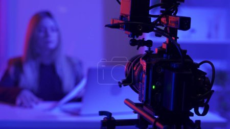 Professional video camera on a tripod against a blurred background of a female presenter in a dark studio illuminated by pink and blue neon lights, close up