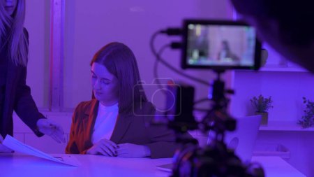 Photo for A female presenter discusses a storyboard with a female producer before filming begins in close up. Women in a dark studio in pink neon light in front of a professional camera - Royalty Free Image
