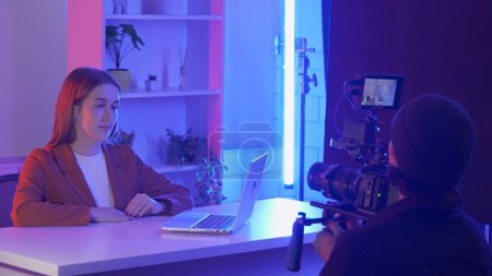 Photo for Backstage video production recording. A videographer uses a professional camera to film a female presenter sitting at a table in front of a laptop. Film crew in the studio in pink and blue neon - Royalty Free Image