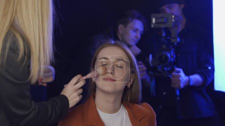 Photo for Crew preparing for shoot in studio. A woman make up artist applies powder to the face of a TV presenter close up. A cinematographer with a camera and an assistant stand waiting in the background - Royalty Free Image