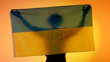 Téléchargez les photos : Drapeaux nationaux concept de publicité patriotique. Silhouette de la personne tenant un grand drapeau sur fond jaune. Silhouette de l'homme avec le drapeau national de l'Ukraine tenant dans les mains. - en image libre de droit
