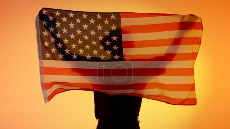 Foto de Banderas nacionales concepto de publicidad patriótica. Silueta de persona sosteniendo gran bandera sobre fondo amarillo. Silueta del hombre con la bandera nacional de los EEUU cogida de la mano. - Imagen libre de derechos