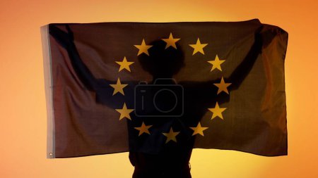 Photo for National flags patriotic advertisement concept. Person silhouette holding big flag against yellow background. Silhouette of man with national flag of European union holding in hands. - Royalty Free Image