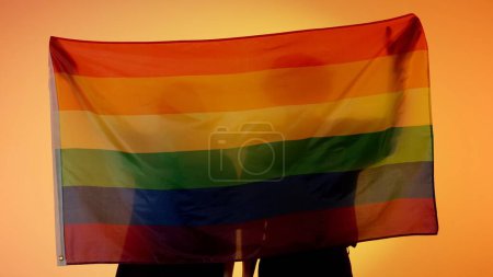 Photo for Love and diversity advertisement concept. Silhouettes of people with large flag on yellow background. Silhouettes of a man and a woman with LGBT pride flag holding in hands. - Royalty Free Image