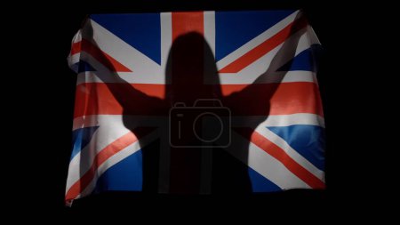 Téléchargez les photos : Drapeaux nationaux concept de publicité patriotique. Silhouette de la personne tenant un grand drapeau sur fond noir. Silhouette de femme avec le drapeau national de la Grande-Bretagne tenant dans les mains. - en image libre de droit