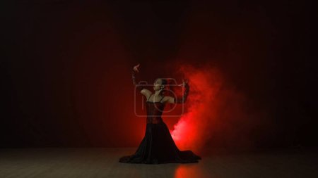 Téléchargez les photos : Chorégraphie moderne une danse. Femme dansant sur fond noir avec lumière rouge et fumée. Une danseuse espagnole en robe rouge et noire montre des éléments de chorégraphie flamenco. - en image libre de droit