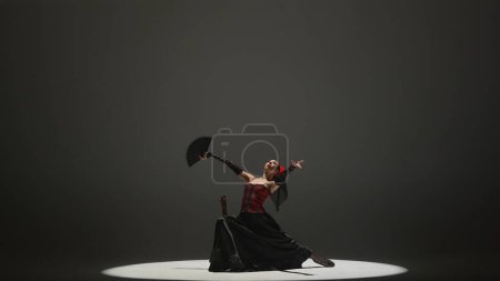 Photo for Modern choreography an dance. Woman dancing on black background under spotlights. Spanish dancer in red-black dress demonstrates elements of flamenco choreography sitting on a chair. - Royalty Free Image