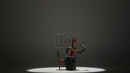 Photo for Modern choreography an dance. Woman dancing on black background under spotlights. Spanish dancer in red-black dress demonstrates elements of flamenco choreography sitting on a chair. - Royalty Free Image