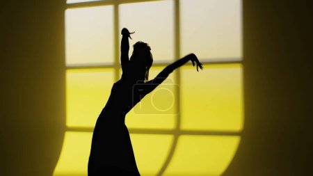 Photo for Modern dance choreography. Female silhouette of dancer dancing against window shade. Woman in flamenco style dress performs elegant Spanish dance movements with hands and body in studio - Royalty Free Image