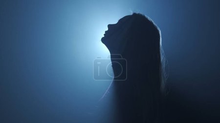 Photo for Silhouette in lighting concept. Female model against bright light on black smoky background in studio. Female silhouette standing surrounded by rays of light in darkness. Side view - Royalty Free Image