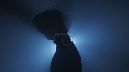 Foto de Silueta en concepto de iluminación. Modelo femenino contra la luz brillante sobre fondo negro ahumado en estudio. Silueta femenina de pie rodeada de rayos de luz en la oscuridad. Vista lateral - Imagen libre de derechos