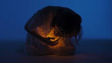 Photo for Contemporary experimental choreography concept. Female dancer performing in the studio. Young woman in dark studio touching warm light glowing inside the chest under her shirt. - Royalty Free Image