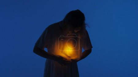 Photo for Contemporary experimental choreography concept. Female dancer performing in the studio. Young woman in dark studio touching warm light glowing inside the chest under her shirt. - Royalty Free Image