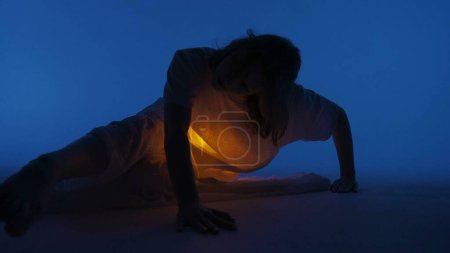 Photo for Contemporary experimental choreography concept. Female dancer performing in the studio. Young woman laying in dark studio with warm light glowing inside the chest under her shirt. - Royalty Free Image
