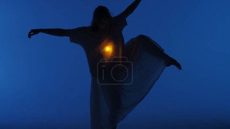 Foto de Concepto contemporáneo de coreografía experimental. Bailarina actuando en el estudio. Mujer joven bailando en estudio oscuro con luz cálida brillando dentro del pecho debajo de su camisa. - Imagen libre de derechos