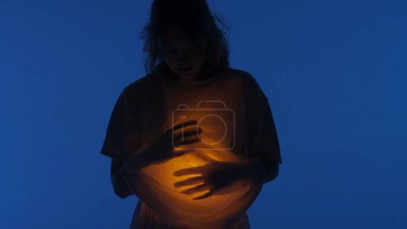 Photo for Contemporary experimental choreography concept. Female dancer performing in the studio. Young woman in dark studio touching warm light glowing inside the chest under her shirt, close up. - Royalty Free Image