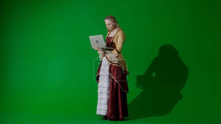 Photo for Historical person modern lifestyle advertisement. Woman in ancient outfit on chroma key green screen background. Female in renaissance style dress holding laptop typing message. - Royalty Free Image