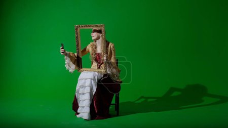 Photo for Historical person modern lifestyle advertisement. Woman in ancient outfit on chroma key green screen background. Female in renaissance style dress holding painting frame taking selfie on smartphone. - Royalty Free Image