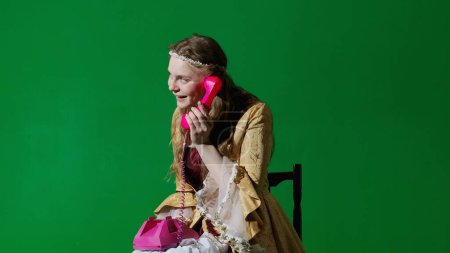 Photo for Historical person modern lifestyle advertisement. Woman in ancient outfit on the chroma key green screen background. Female in renaissance style dress holding old retro phone calling and talking. - Royalty Free Image