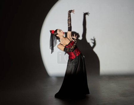 Photo for Modern choreography an dance. Woman dancing on black background under spotlights. Spanish dancer in red-black dress dancing elements of passionate flamenco. - Royalty Free Image