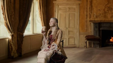 Photo for Historical person modern lifestyle advertisement. Woman in ancient outfit on background of historic interior. Female in renaissance style dress sitting drinking coffee from paper cup. - Royalty Free Image