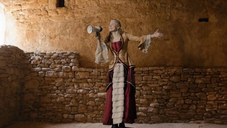 Historische Person moderne Lifestyle-Werbung. Frau im antiken Outfit vor dem Hintergrund des historischen Interieurs. Frau im Renaissance-Kleid hält Megafon in der Hand.