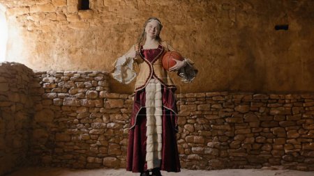 Photo for Historical person modern lifestyle advertisement. Woman in ancient outfit on background of historic interior. Female in renaissance style dress holding basketball ball smiles shows thumb up. - Royalty Free Image