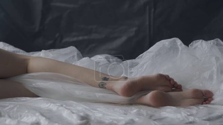 Körperschönheit und Modewerbung. Weibchen liegen auf dem Bett im Studio. Beine einer jungen Frau in Nahaufnahme, die unter einer dünnen Folie aus Wachstuch liegt.