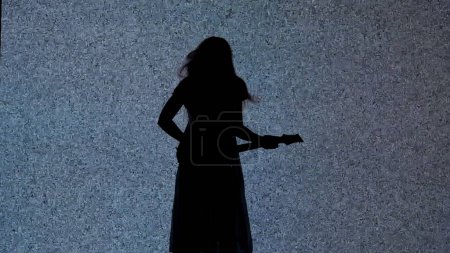 Photo for Halloween horror movie creative concept. Silhouette against digital television screen. Thriller scene woman silhouette in dress playing on guitar in front of big digital screen with white noise. - Royalty Free Image