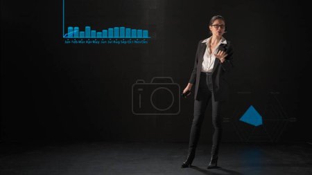 Photo for Business conference presentation lecture. Woman presenting project on stage during meeting. A businesswoman talks to the audience, shows infographics, revenue analysis on a huge LED screen. - Royalty Free Image