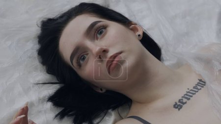 Beauté corporelle et publicité de mode. Femme en sous-vêtements allongée sur le lit dans le studio. Jeune femme seminude en lingerie pose sous une mince feuille de tissu huilé envelopper, fixer la caméra.