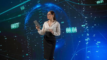 Foto de Una mujer en un traje de negocios da una conferencia mientras usa una tableta para presentar datos complejos en una pantalla digital grande. Espacio digital, un concepto espacial. - Imagen libre de derechos