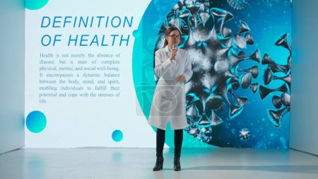 A professional woman in a white lab coat stands in front of a drug product on a large digital screen giving a scientific lecture. The concept of medicine, health and health care.