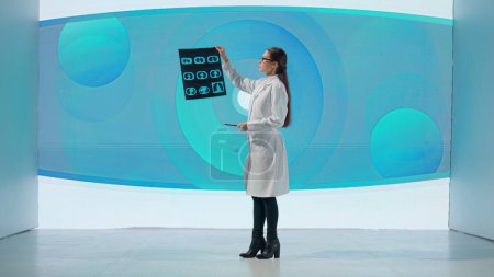 Foto de Una mujer con una bata blanca se para frente a un producto de drogas en una gran pantalla digital. Una científica muestra una imagen de los pulmones y da una conferencia científica - Imagen libre de derechos