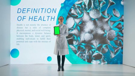 Foto de Una mujer con una bata blanca se para frente a un producto de drogas en una gran pantalla digital. Una científica demuestra una tableta con una pantalla verde y da una conferencia científica - Imagen libre de derechos