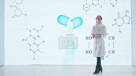 Téléchargez les photos : Une femme professionnelle en blouse blanche se tient devant un produit pharmaceutique sur un grand écran numérique donnant une conférence scientifique. Le concept de médecine, de santé et de soins de santé. - en image libre de droit