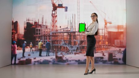 Una mujer interactúa con una proyección de un edificio de varios pisos y muestra una tableta de pantalla verde. La pantalla digital ilumina el espacio con la estética de un plano.