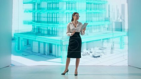 A woman interacts with a holographic projection of a multi-story building. The digital display illuminates the space with the aesthetics of a blueprint, symbolizing advanced architectural design