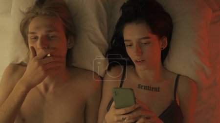 Photo for Love intimate relationships concept. Top view loving couple romantic time in bedroom. Man and woman sensual lovers laying in bed together, female texting on smartphone, male smoking cigarette. - Royalty Free Image