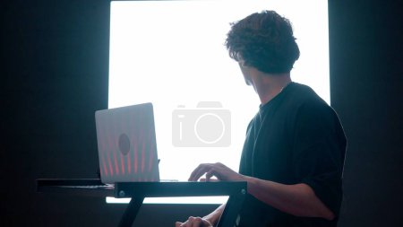 Photo for Digital visual technology concept. Young male working on laptop against digital wall in dark club. Man silhouette sitting typing on laptop, gaming mixing music in front of big digital screen. - Royalty Free Image