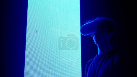 Photo for Digital visual technology concept. Man silhouette in virtual reality glasses standing in front of digital screen wall with neon symbols looking around playing video game. - Royalty Free Image