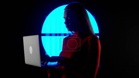 Téléchargez les photos : Concept de technologie visuelle numérique. Silhouette féminine avec ordinateur portable contre le mur numérique dans le club sombre. Femme tapant sur ordinateur portable, jeu mélangeant la musique devant l'écran numérique avec des symboles.. - en image libre de droit