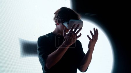 Téléchargez les photos : Concept de technologie visuelle numérique. Silhouette d'homme dans des lunettes de réalité virtuelle debout devant le mur d'écran numérique avec des symboles abstraits regardant autour tenant la main vers le haut. - en image libre de droit