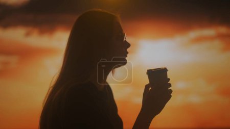 Foto de Silueta de una joven hembra bebiendo café de una taza de papel y disfrutando del aroma en el fondo de los colores brillantes de la puesta del sol. - Imagen libre de derechos