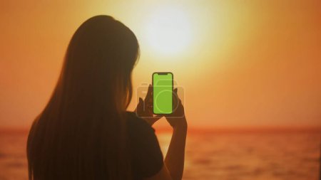 Foto de Silueta de una joven hembra con un smartphone en sus manos buscando información, sobre un fondo de tonos brillantes de puesta de sol. - Imagen libre de derechos